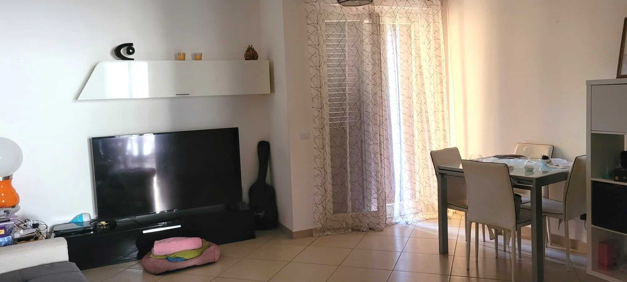 Interno di un soggiorno luminoso con TV, divano e zona pranzo, arredato in stile moderno con tende leggere e accessori decorativi.