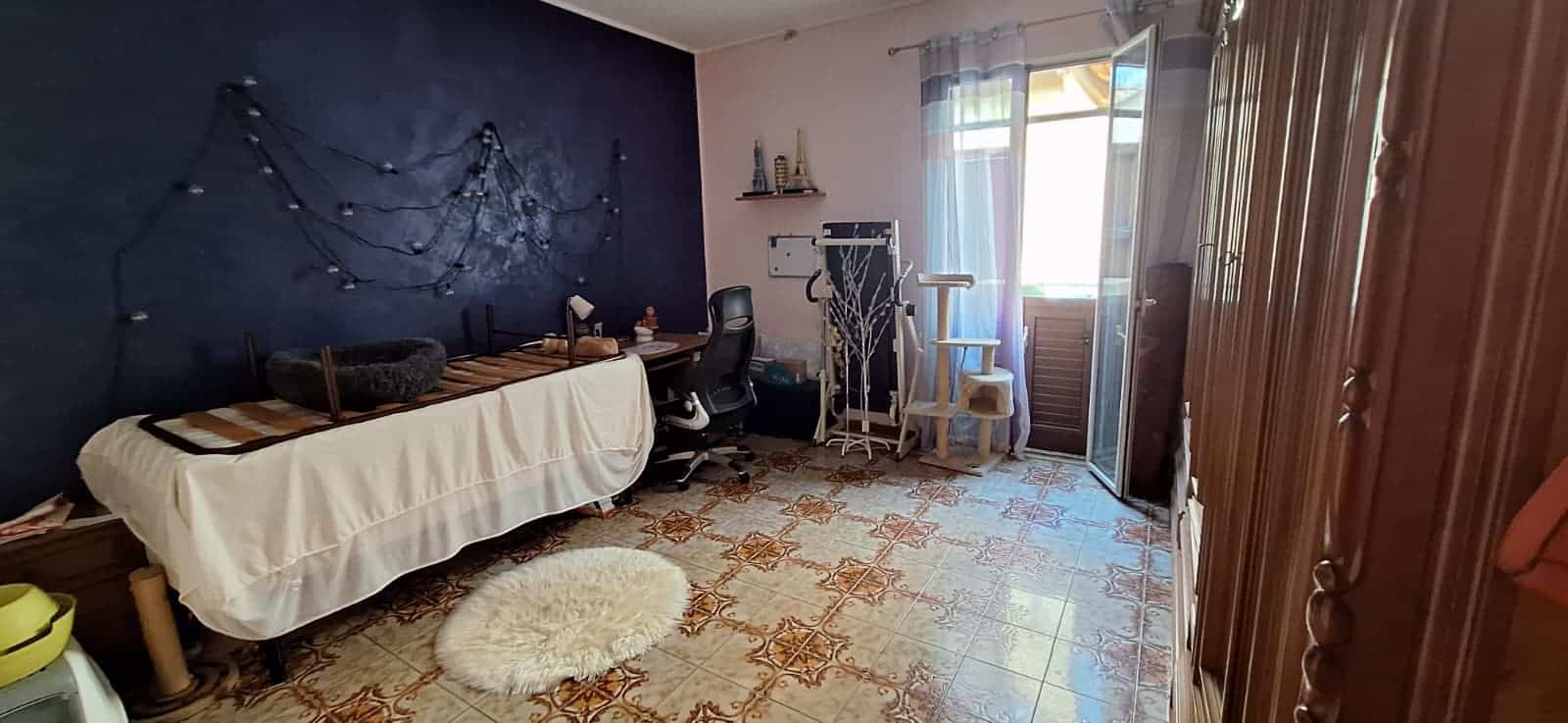 Spazio interno di una stanza con pareti blu, decorazioni luminose, scrivania, sedia, e accessori vari, con porta finestra che conduce a un balcone, illuminata dalla luce naturale.