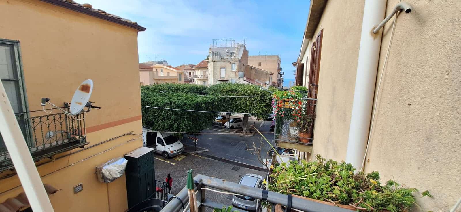 Vista urbana con edifici residenziali, strada e veicoli, cielo sereno, inquadratura da un balcone o finestra, atmosfera tranquilla e quotidiana.