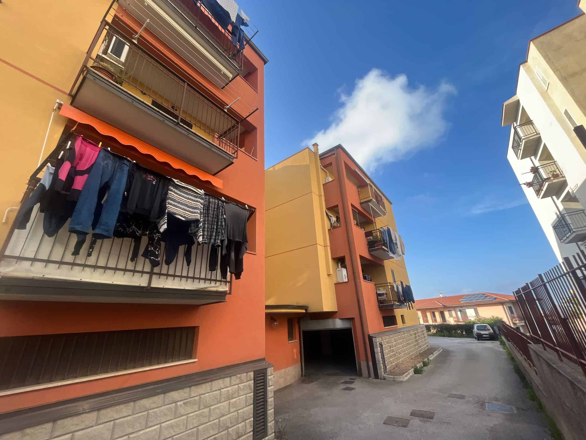 Lavatrice e vestiti stesi su un balcone di un appartamento in un complesso residenziale, con edifici colorati e un cielo sereno.