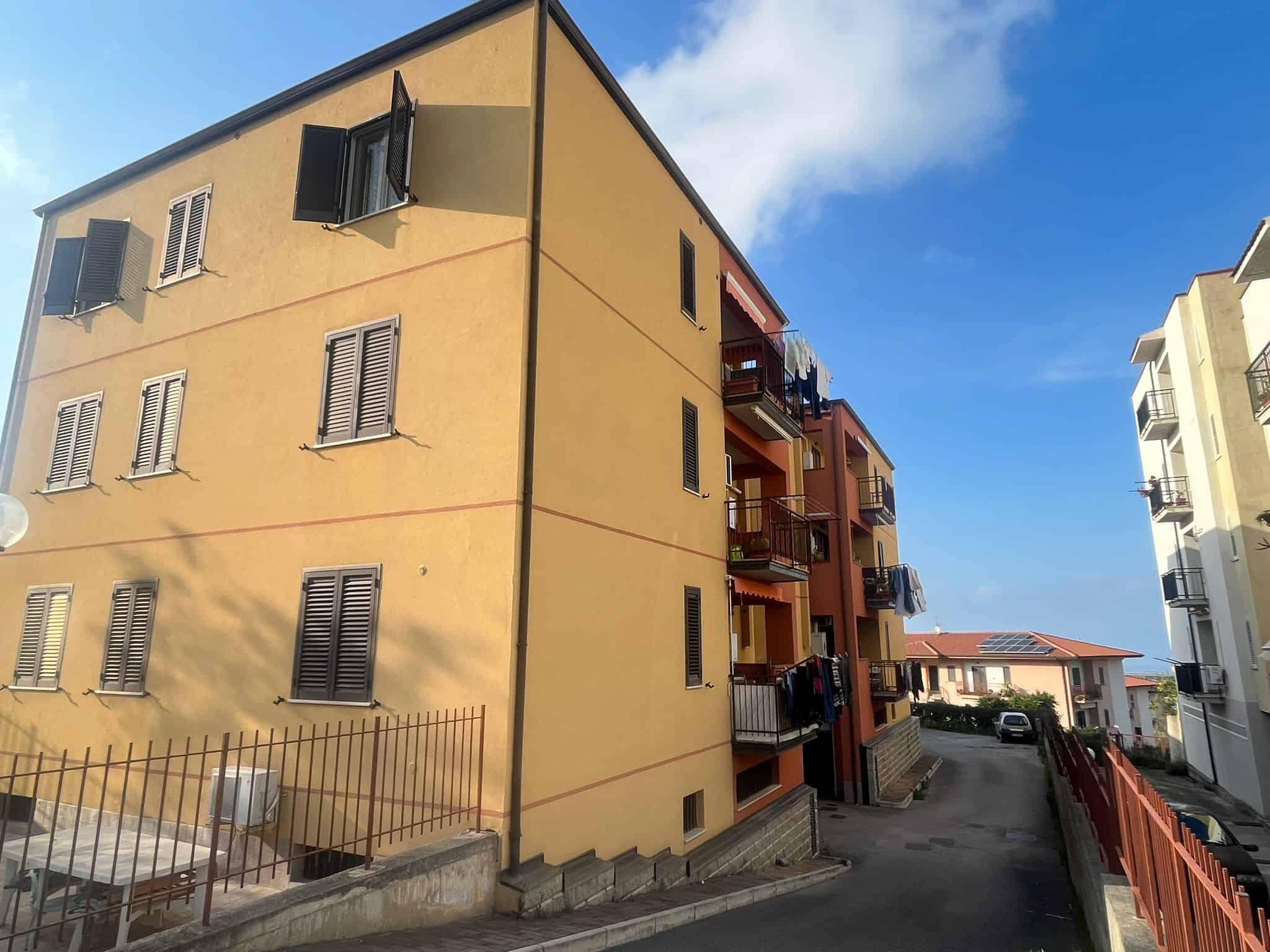 Appartamento in edificio residenziale con balconi e finestre con persiane, situato in zona urbana soleggiata e tranquilla, con strada asfaltata e spazi esterni ben curati, ideale per famiglie o invest.