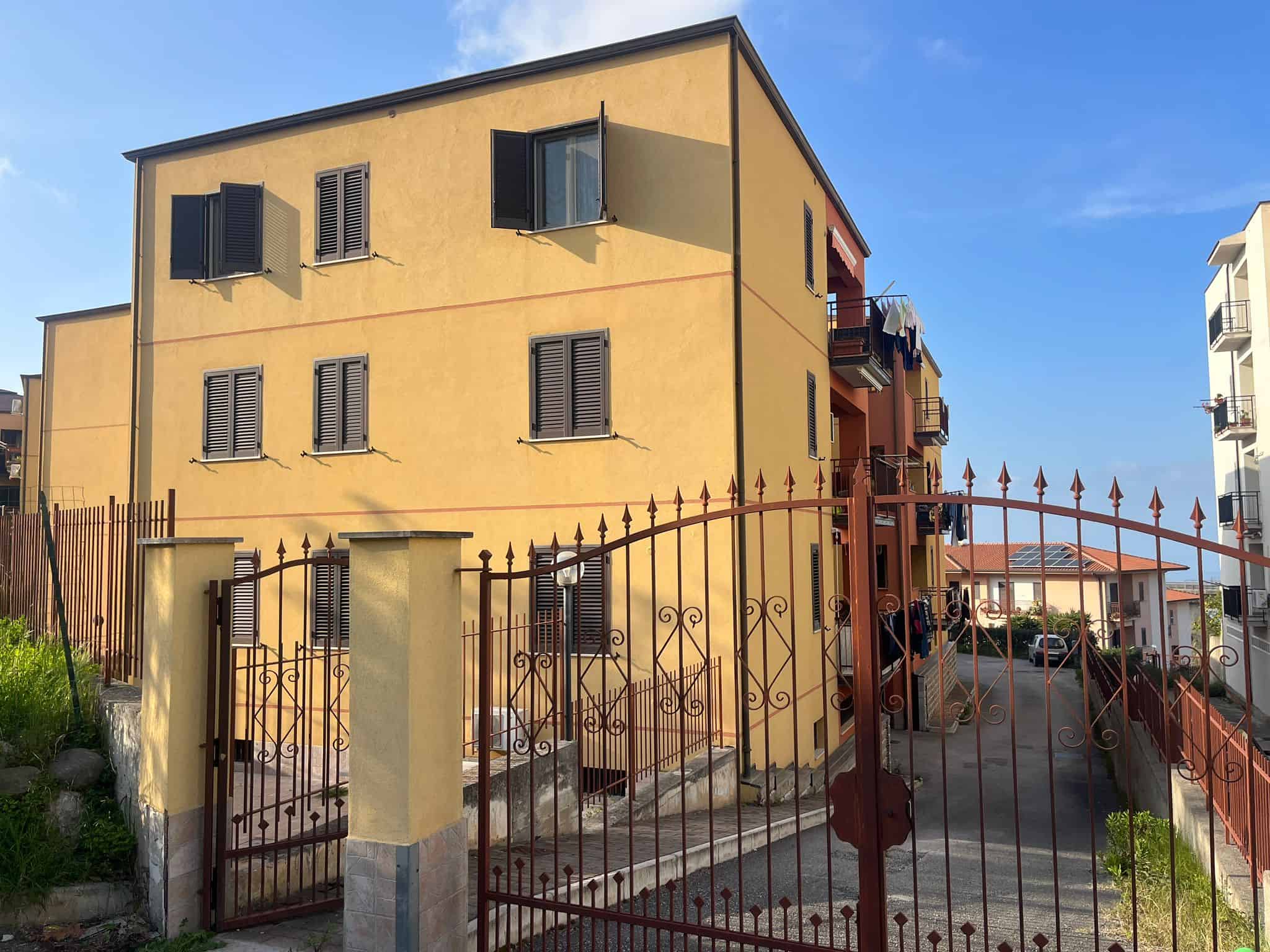 Edificio residenziale con facciata gialla, finestre con persiane nere e cancello in ferro battuto, situato in zona urbana.