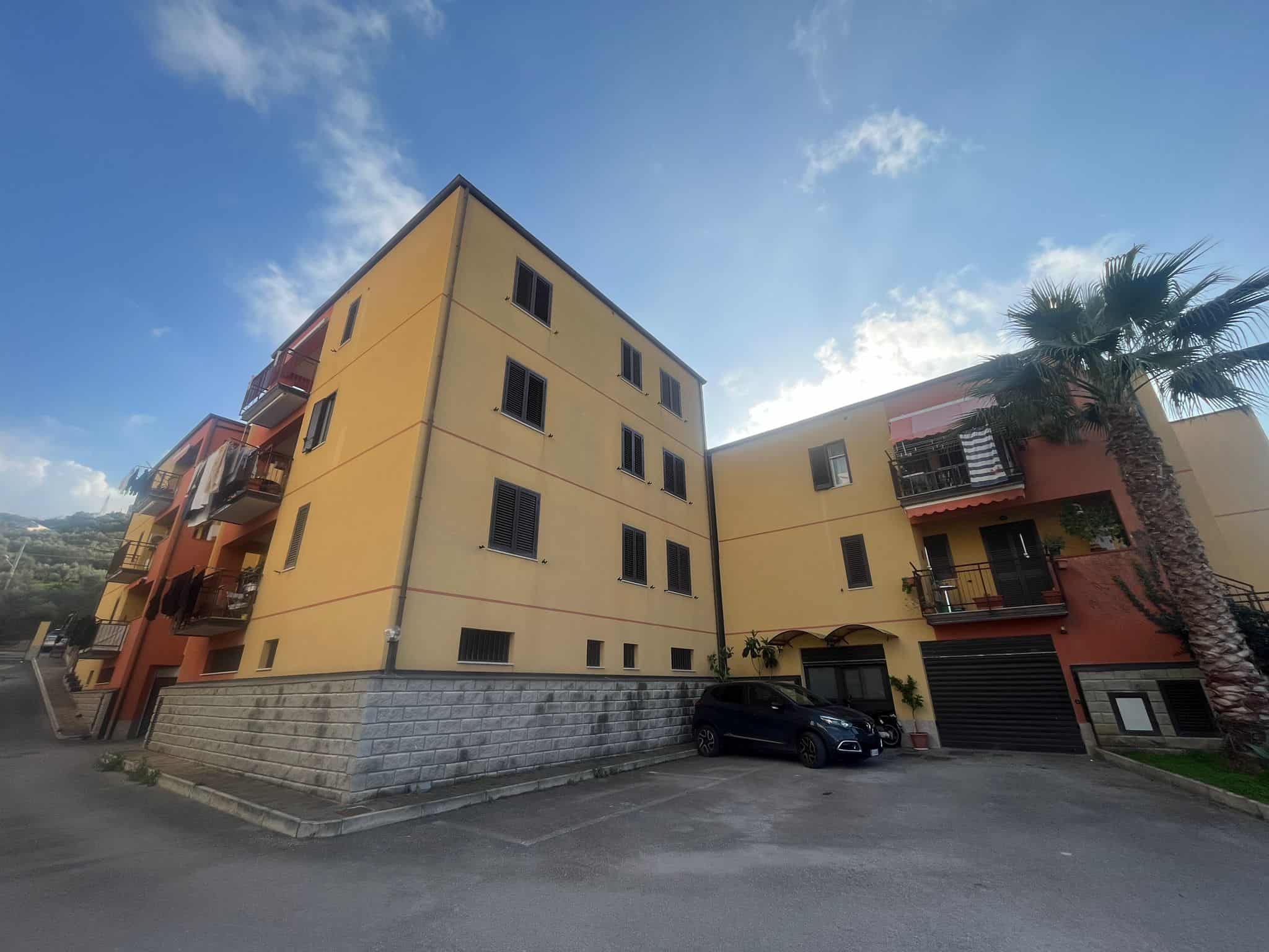 Appartamento residenziale moderno in edificio a più piani con balconi e garage, situato in zona urbana con palmizi e strada asfaltata, sotto un cielo sereno con alcune nuvole.