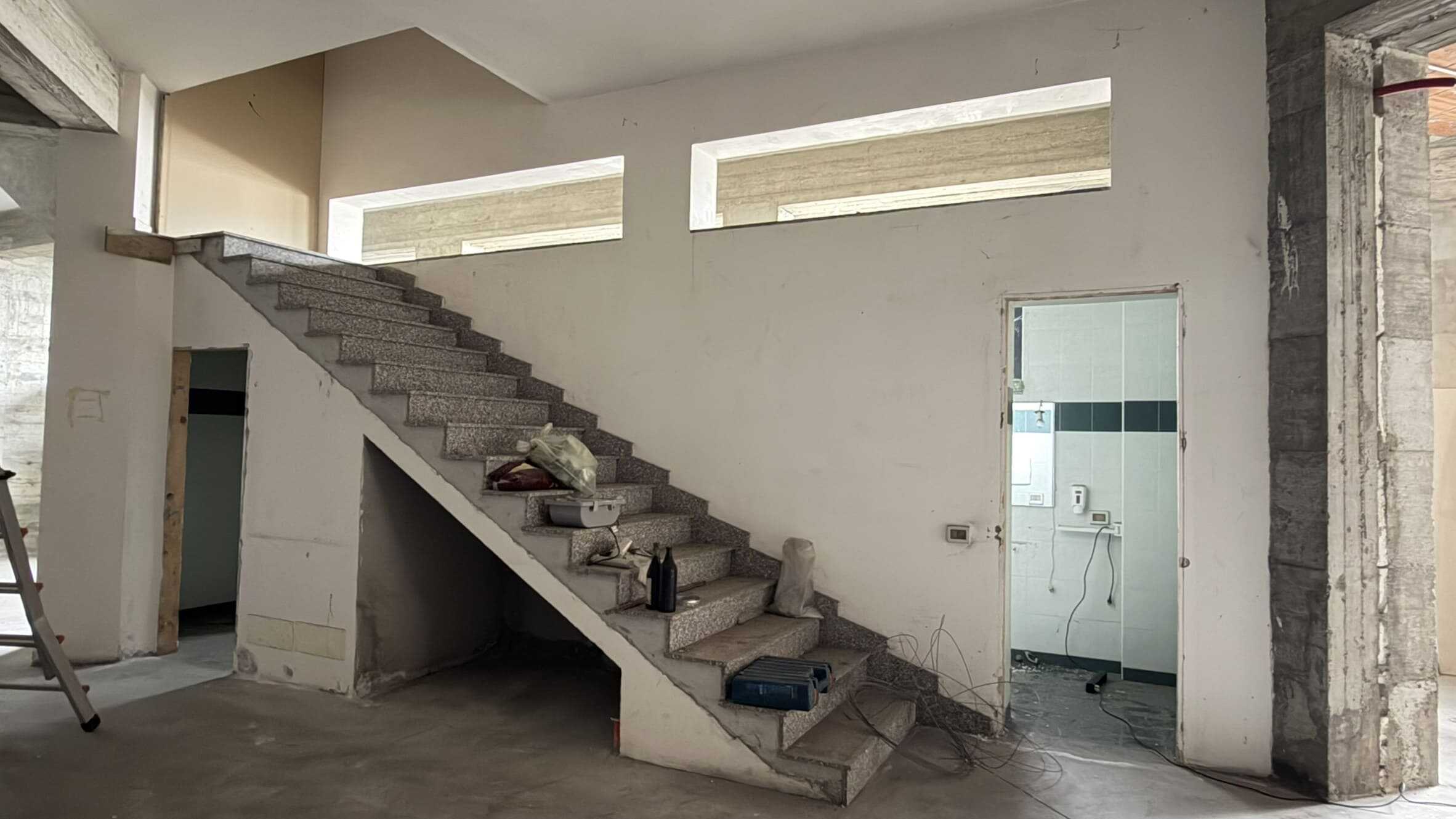 Escalier in cemento in un ambiente di costruzione o ristrutturazione, con pareti bianche e finestre alte che lasciano entrare luce naturale.