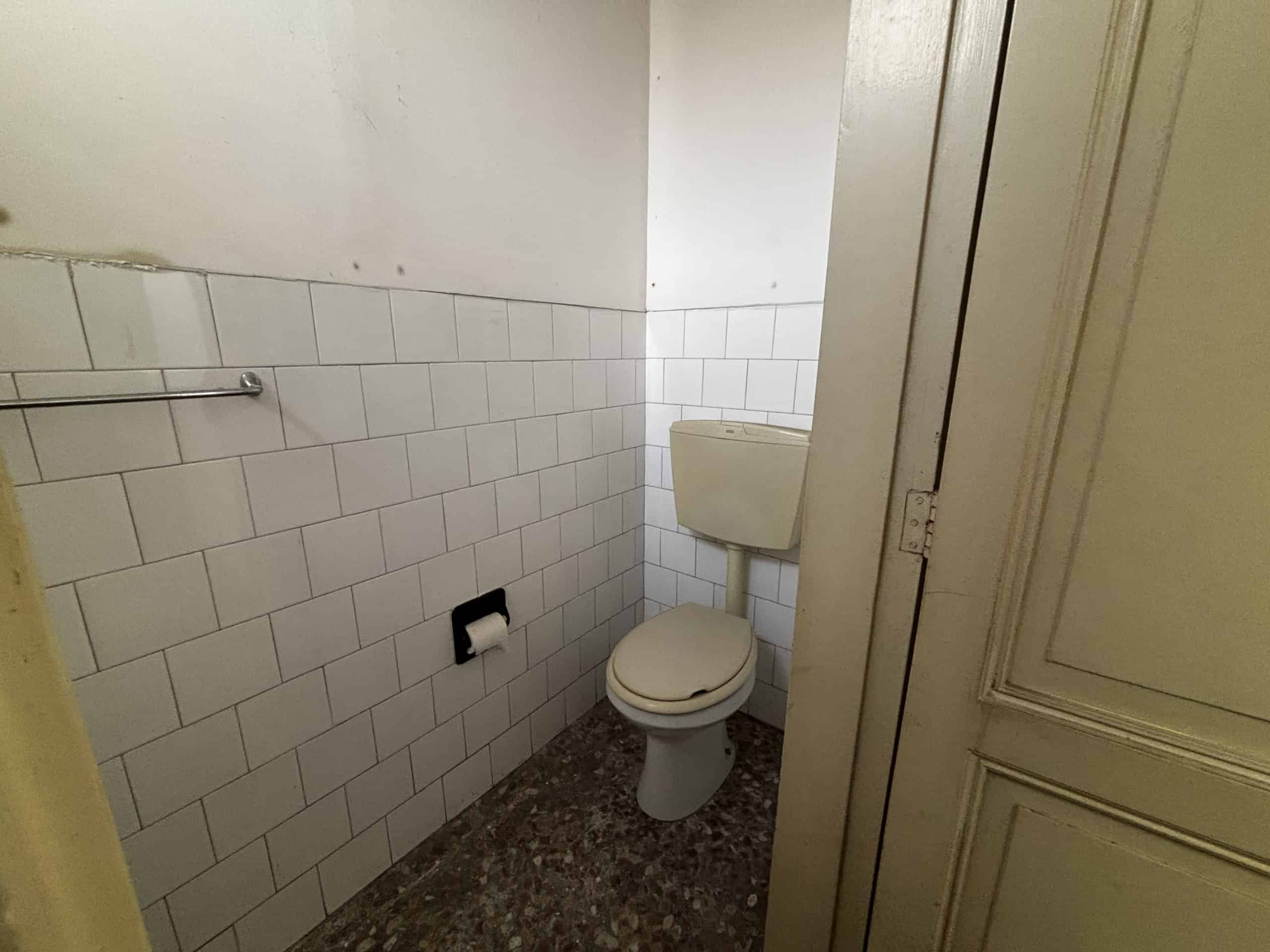 Il bagno con WC e pareti rivestite in piastrelle bianche, pavimento in marmo scuro, in un ambiente compatto e funzionale.