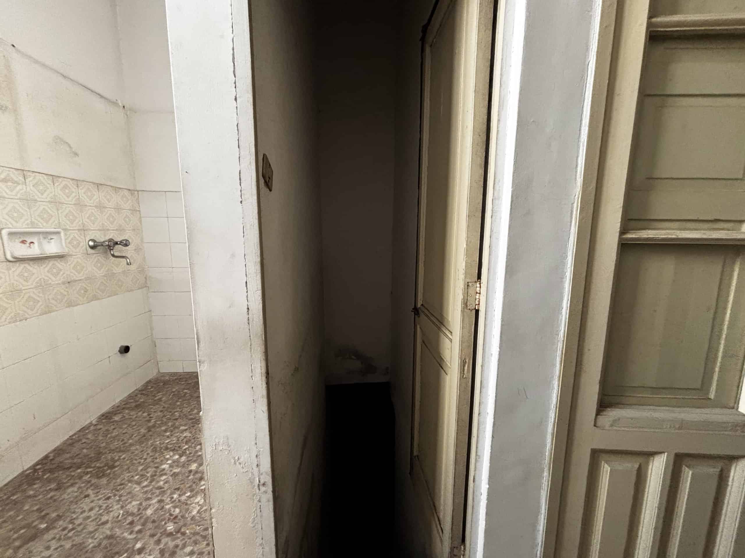 Scorcio di un angolo di cucina con pareti bianche e superficie in granito, mostra un ingresso stretto e buio verso un'altra stanza, evidenziando uno spazio abitativo datato e da ristrutturare.