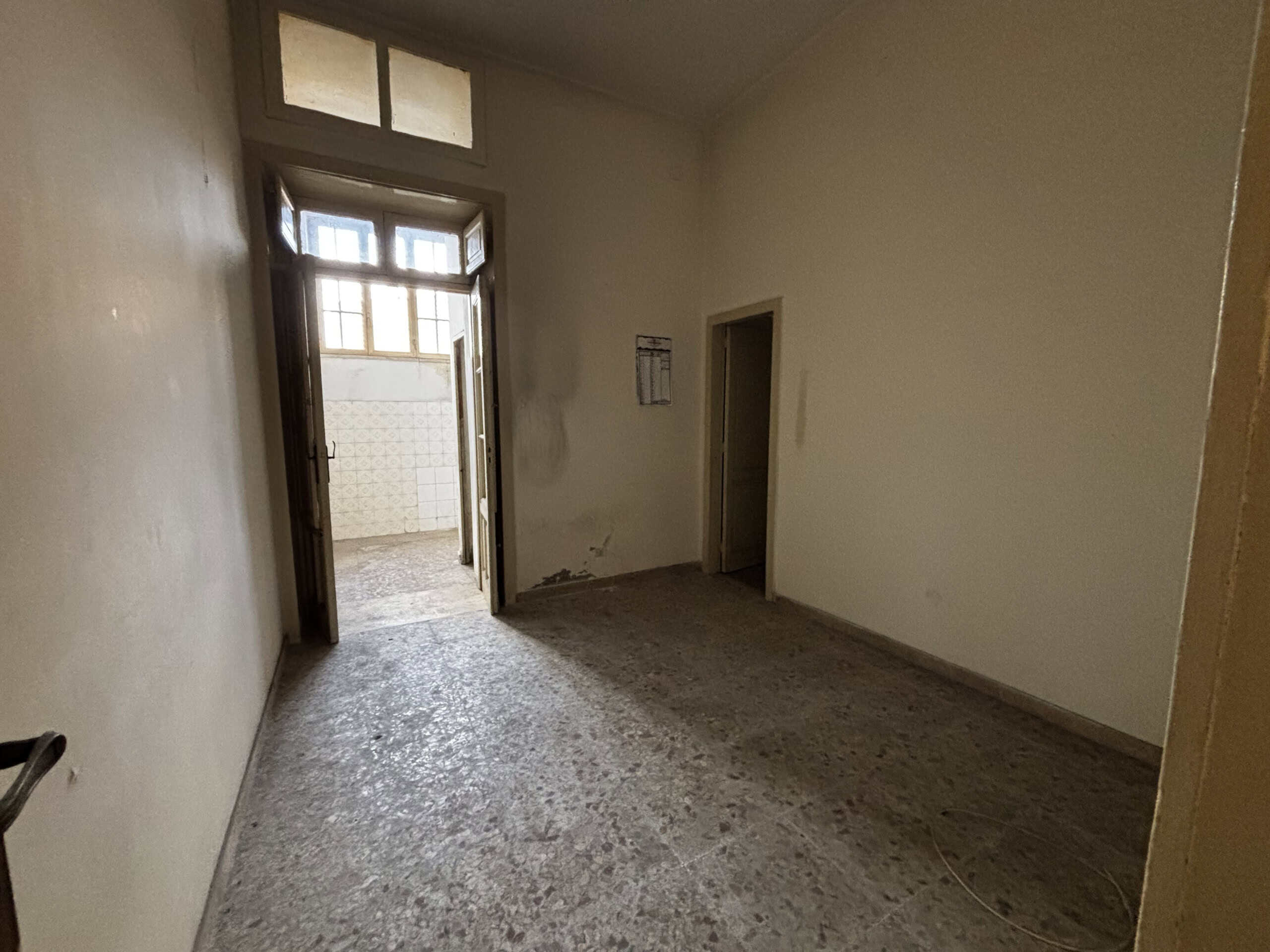 Interno di un appartamento vuoto con pareti bianche, pavimento in cemento e porta finestra che conduce a un balcone, in condizioni da ristrutturare.
