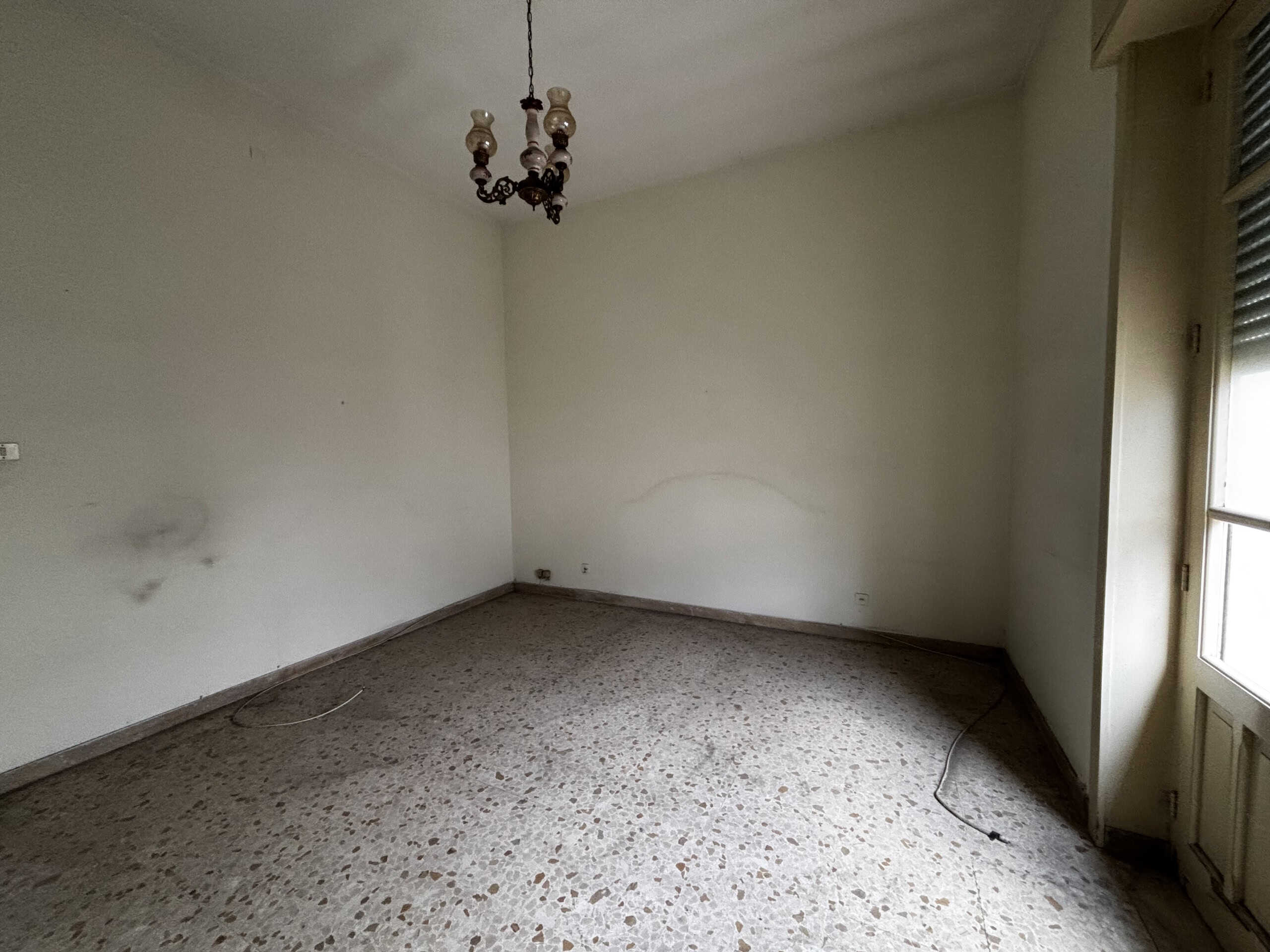 Appartamento vuoto con pareti bianche, pavimento in marmo e lampadario antico, luce naturale da finestra, ideale per ristrutturazione e personalizzazione.