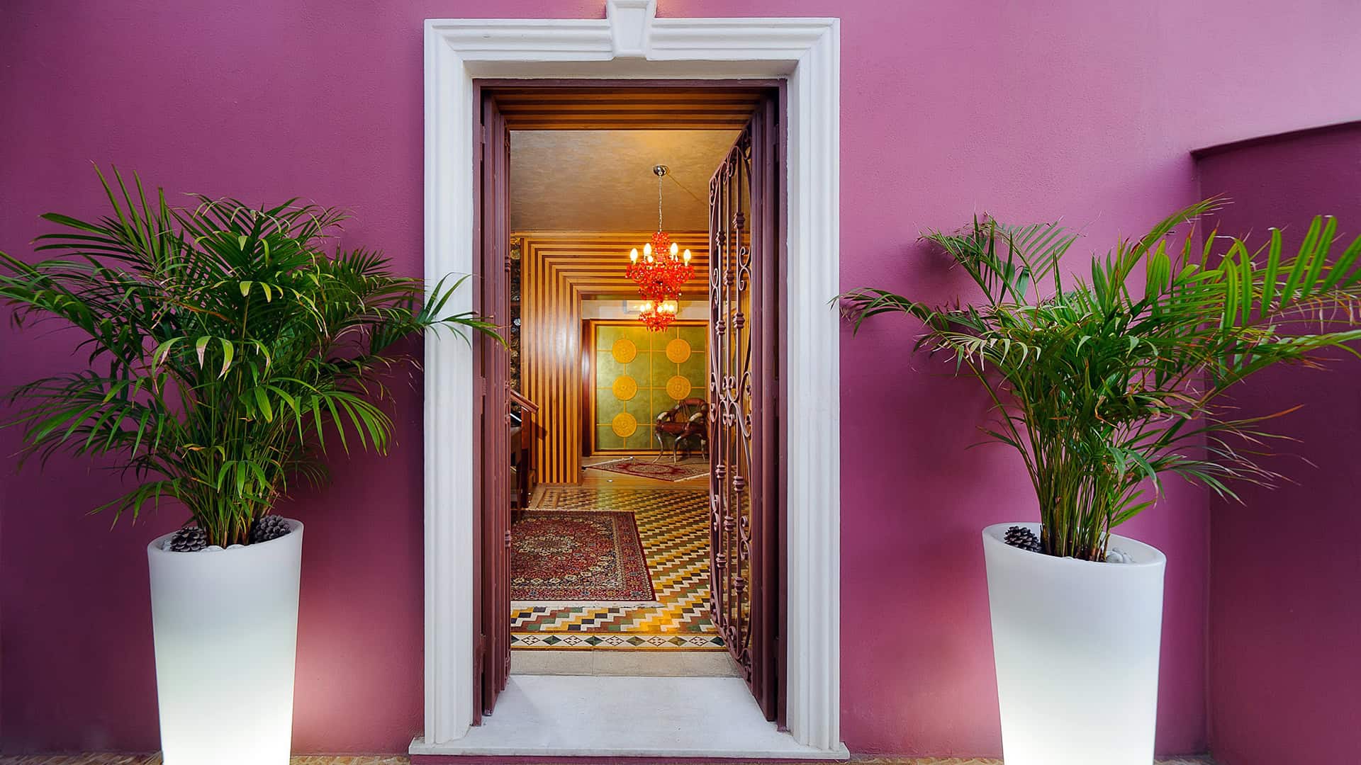 Ingresso di una casa elegante con pareti rosa, piante verdi in vasi bianchi e porta aperta che rivela un interno decorato con mobili di design e dettagli colorati.