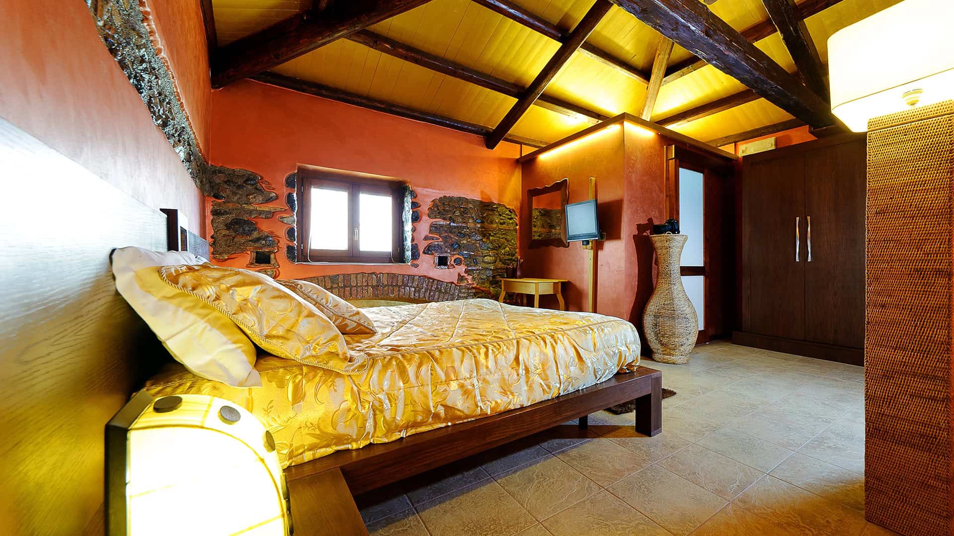 Letto matrimoniale in una camera da letto accogliente con pareti color terracotta, soffitto a travi in legno e grandi finestre che lasciano entrare luce naturale, arredamento caldo e rustico.