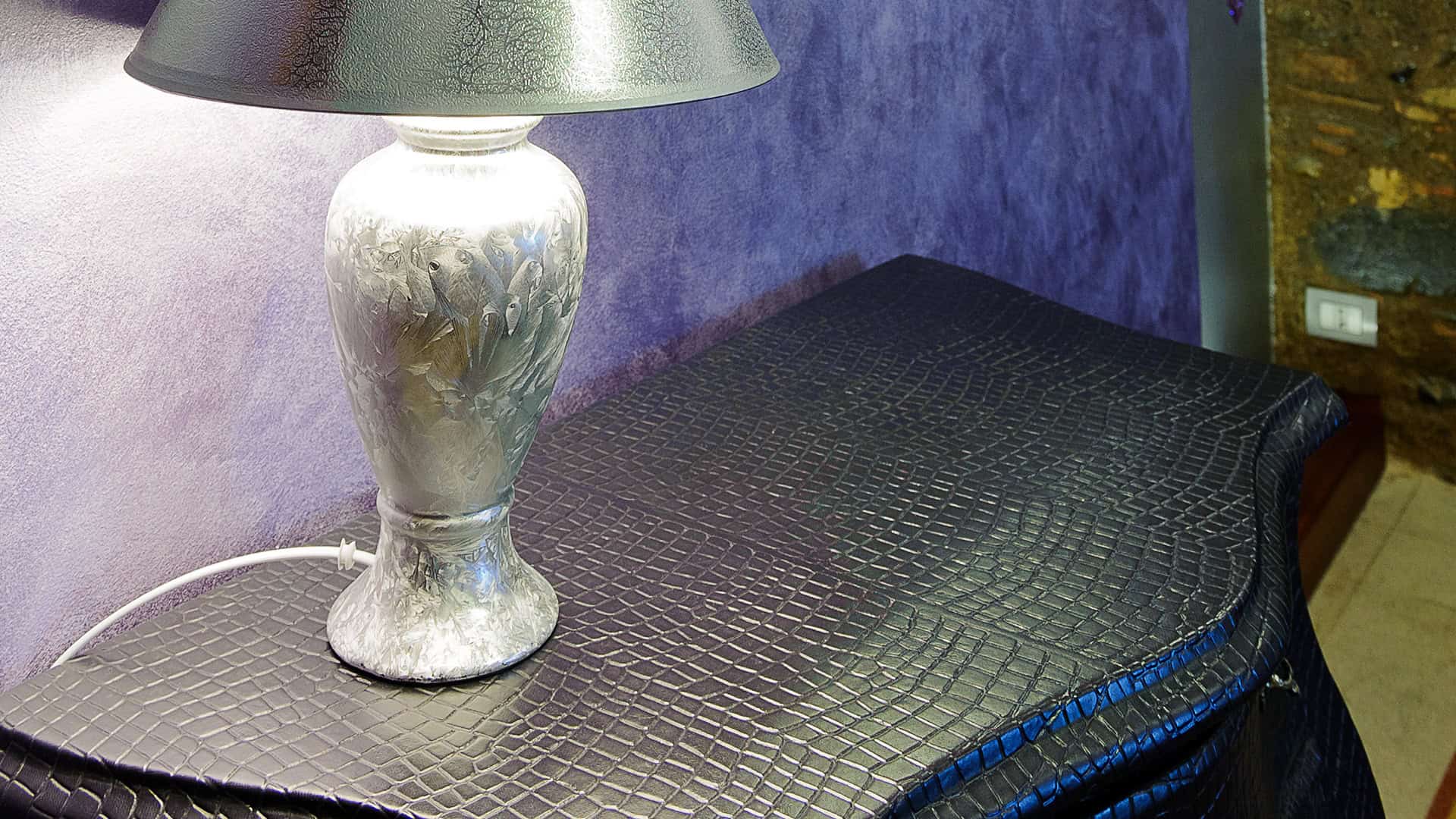 Elegante lampada da tavolo con base in ceramica decorata, posizionata su un mobile nero con superficie a effetto pelle, in un ambiente moderno e raffinato.