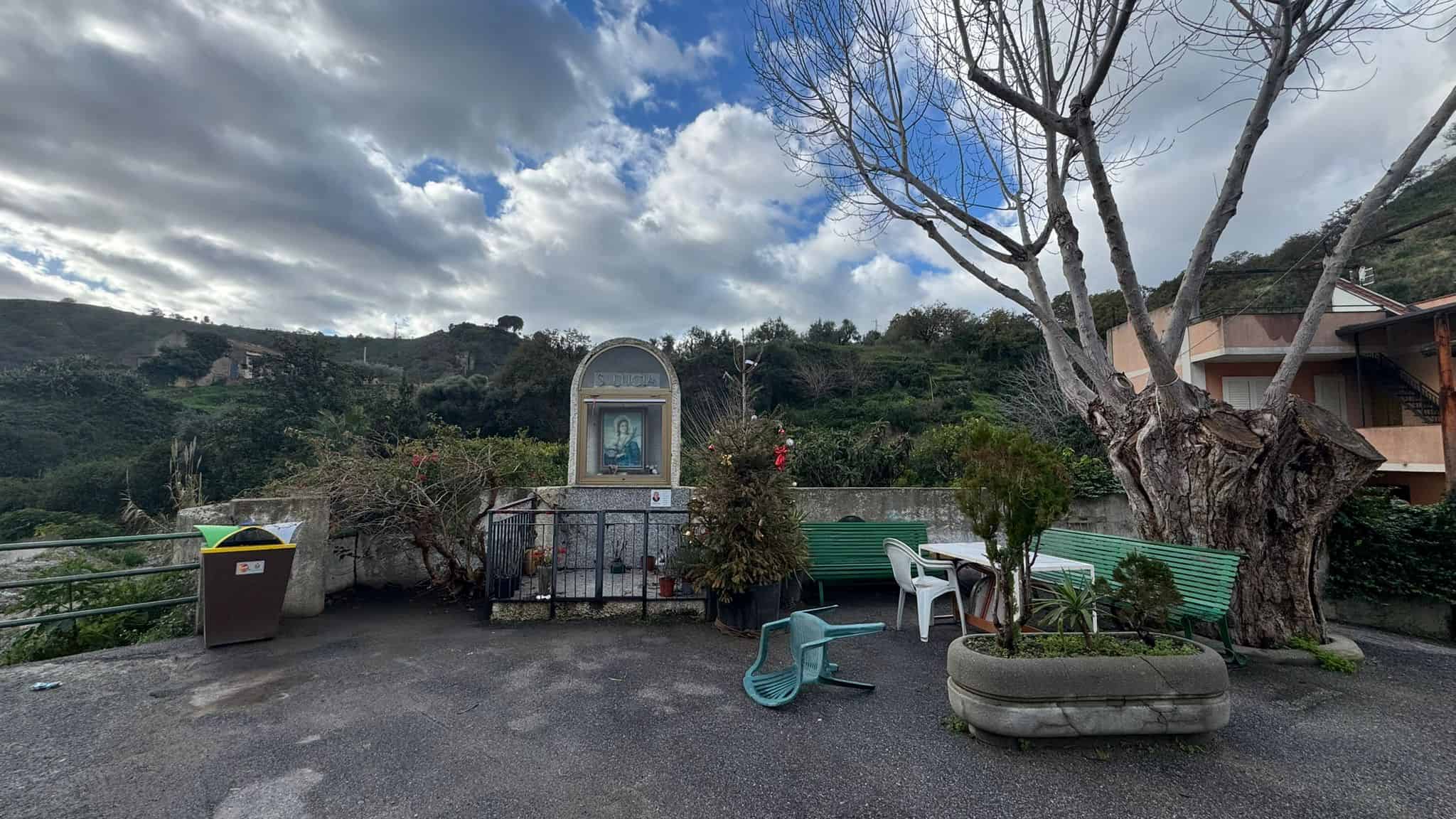 Piazza all'aperto con panchine, alberi e un'edicola religiosa, immersa in un paesaggio collinare con cielo nuvoloso, ideale per momenti di relax e contemplazione all'aperto.
