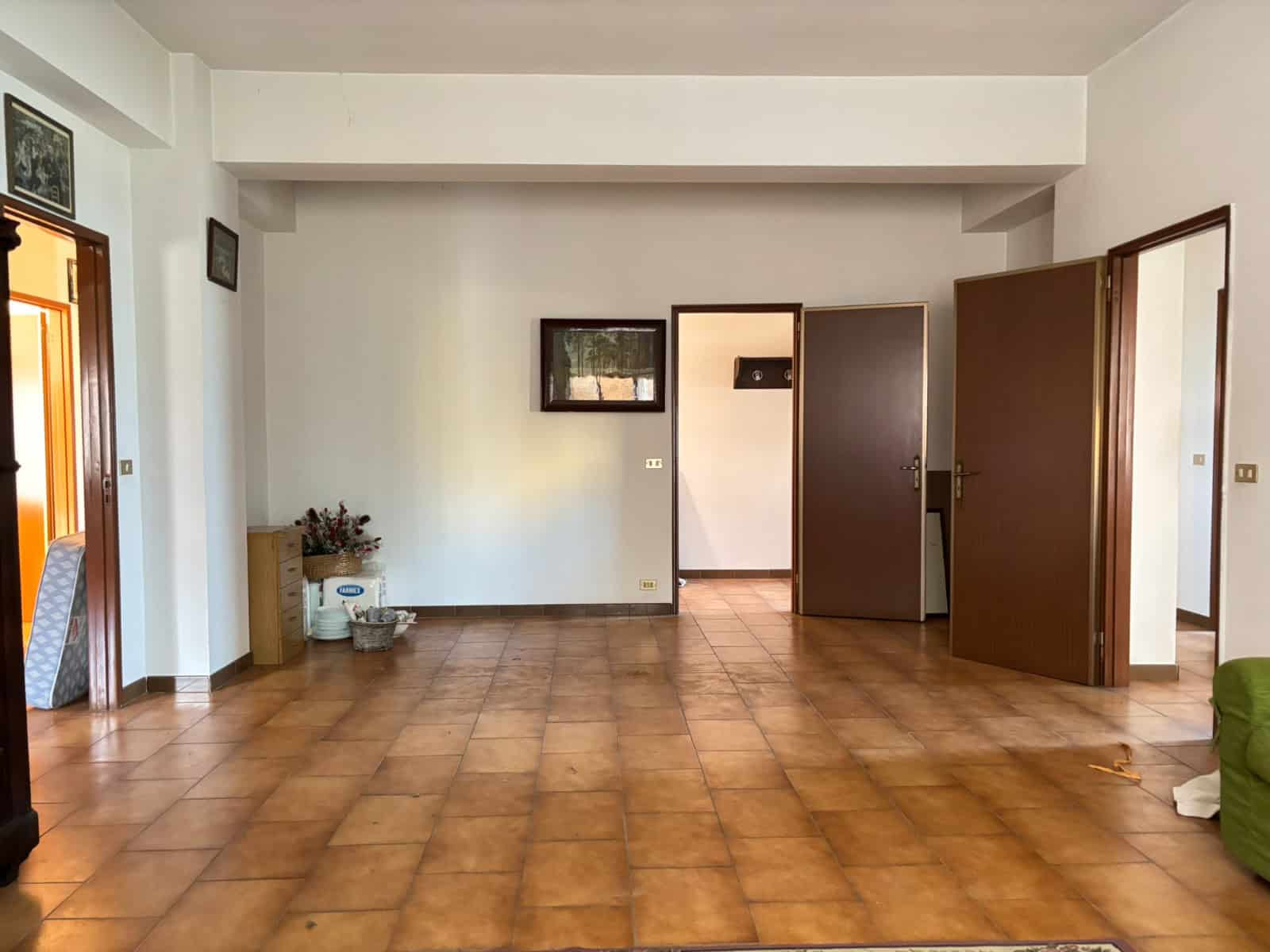 Appartamento vuoto con pareti bianche, porte in legno e pavimento in piastrelle marroni, vista di un soggiorno spazioso e luminoso con ingresso a altre stanze.