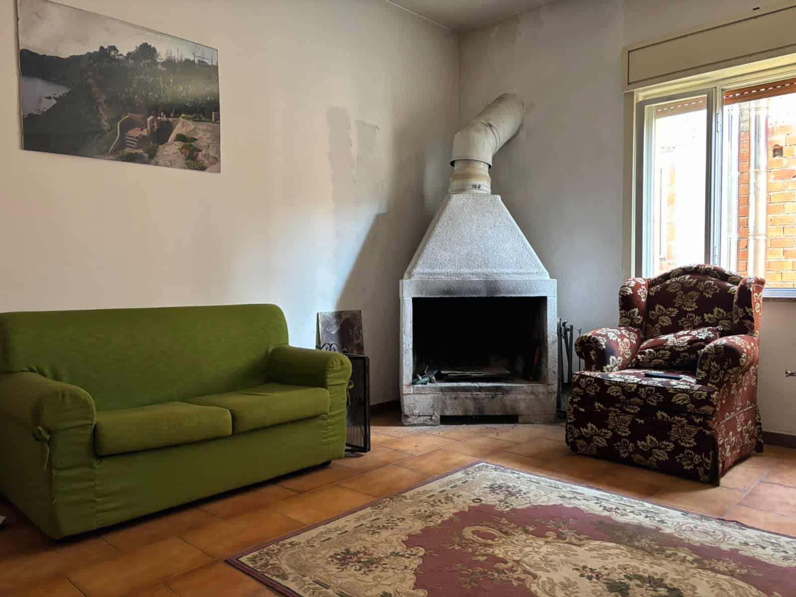 Soggiorno con divano verde, poltrona imbottita con rivestimento floreale, camino in muratura e grande finestra che lascia entrare luce naturale, arredamento accogliente e stile rustico.