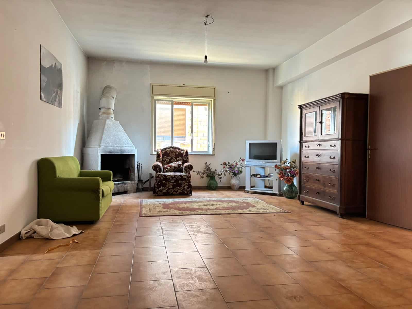 Interno soggiorno con camino, poltrona floreale, divano verde, armadio in legno e TV, ambiente luminoso e accogliente con pavimento in piastrelle e finestra che lascia entrare luce naturale.
