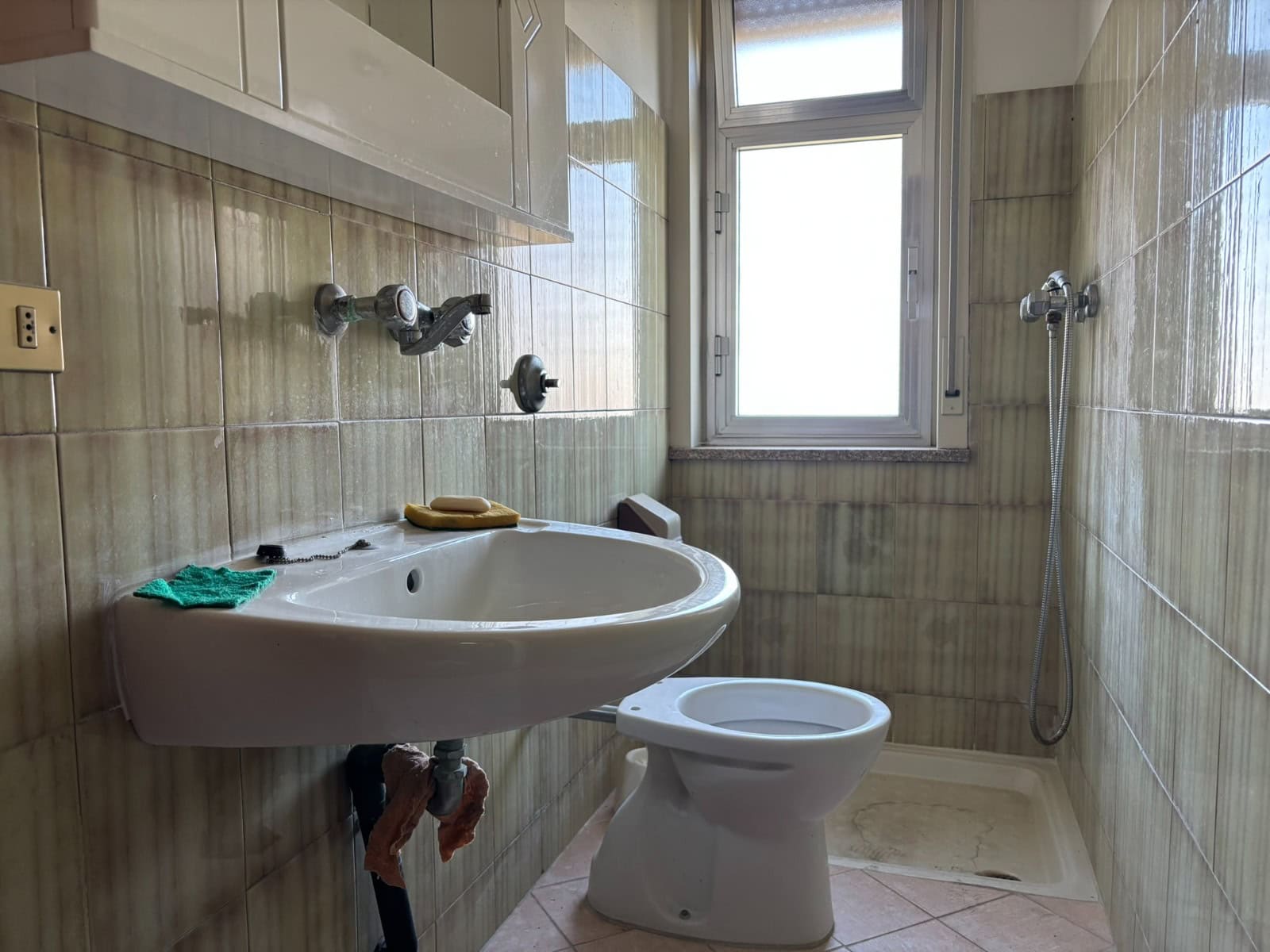 Lavabo e servizi igienici in un bagno di piccole dimensioni con pareti rivestite in piastrelle beige, finestra per ventilazione naturale e doccia a parete, ideale per appartamenti moderni.