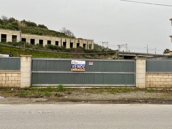 Cancello in metallo con segnale di vendita, situato lungo una strada in un'area residenziale in costruzione, con edifici e terreni verdi sullo sfondo.