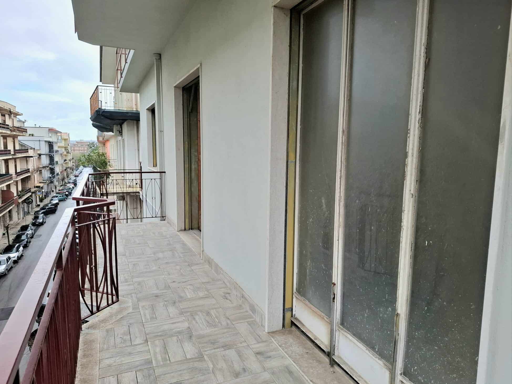 Balcone di un appartamento in città con vista su edifici e strada, pavimentato in piastrelle, con ringhiera in metallo e porta finestra in vetro, in un contesto urbano.