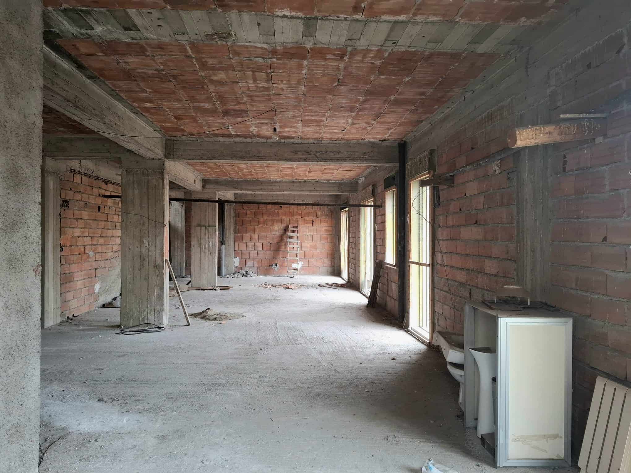 Ampia stanza in costruzione con pareti in mattoni e soffitto in cemento, illuminata da grandi finestre, pronta per essere rifinita e personalizzata.