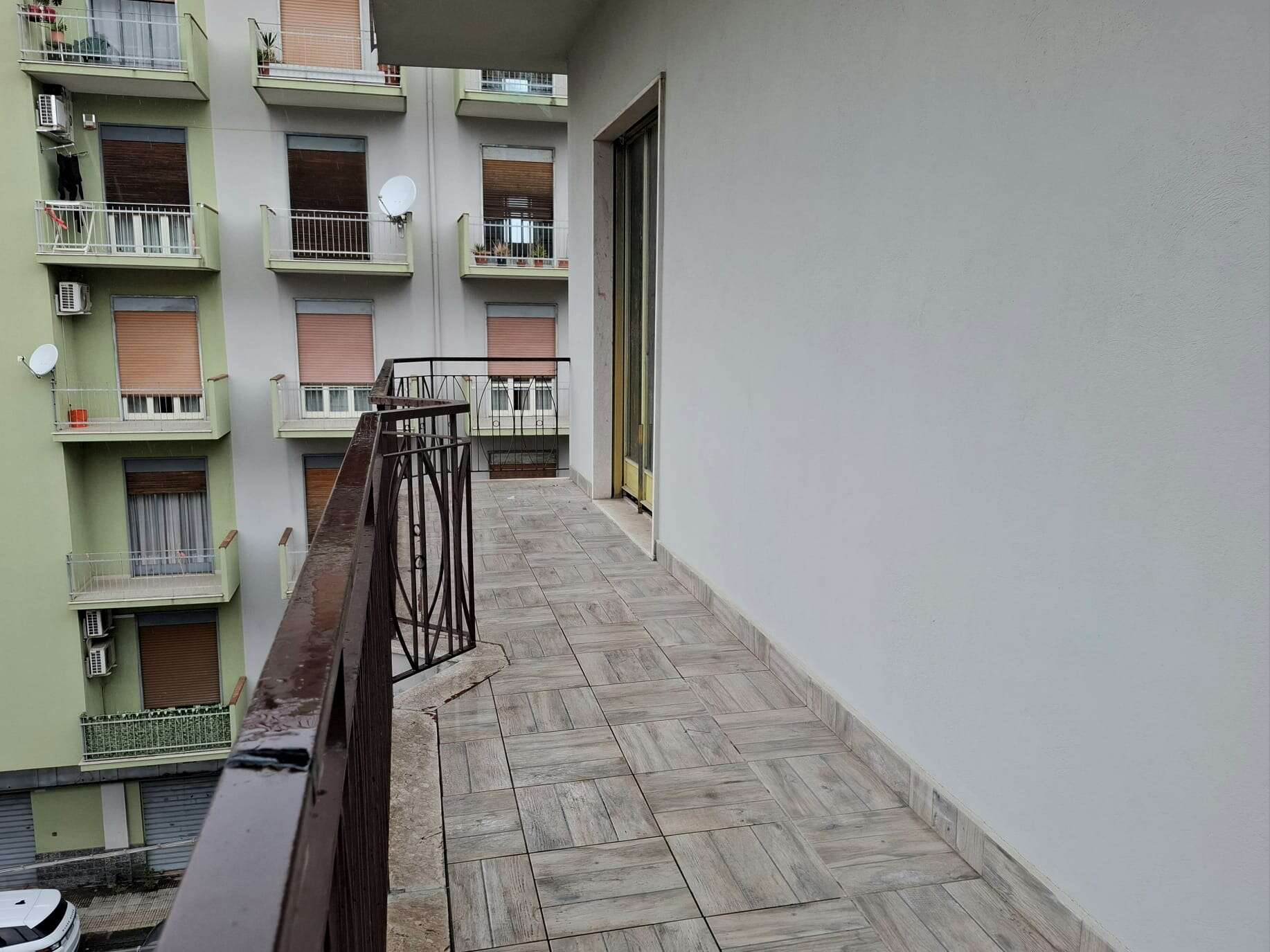 Balcone di un appartamento in un edificio residenziale con vista su altre unità abitative, pavimentato in piastrelle e con ringhiera in metallo, in un contesto urbano con più balconi e finestre.