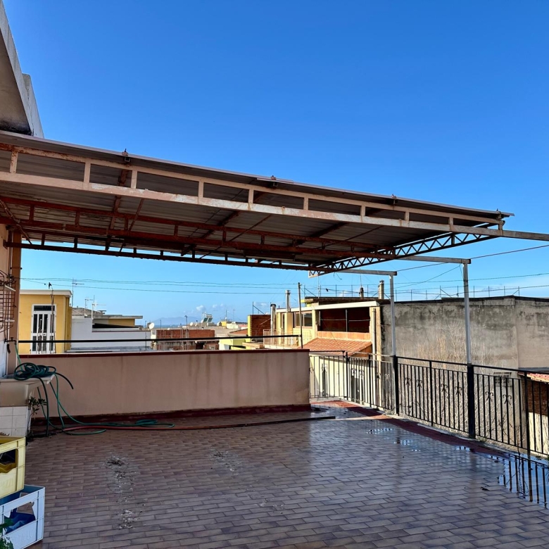Tettoia in metallo e copertura in vetro su un ampio terrazzo urbano con vista su edifici residenziali, ideale per spazi esterni e relax all'aperto.