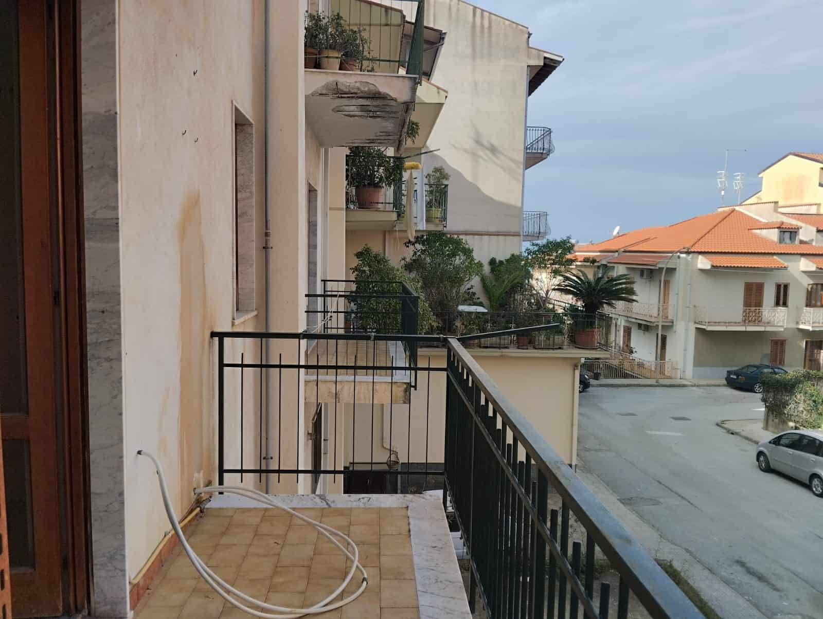 Balcone di un appartamento con vista su strada e altre abitazioni, con ringhiera in metallo e piante sul balcone.