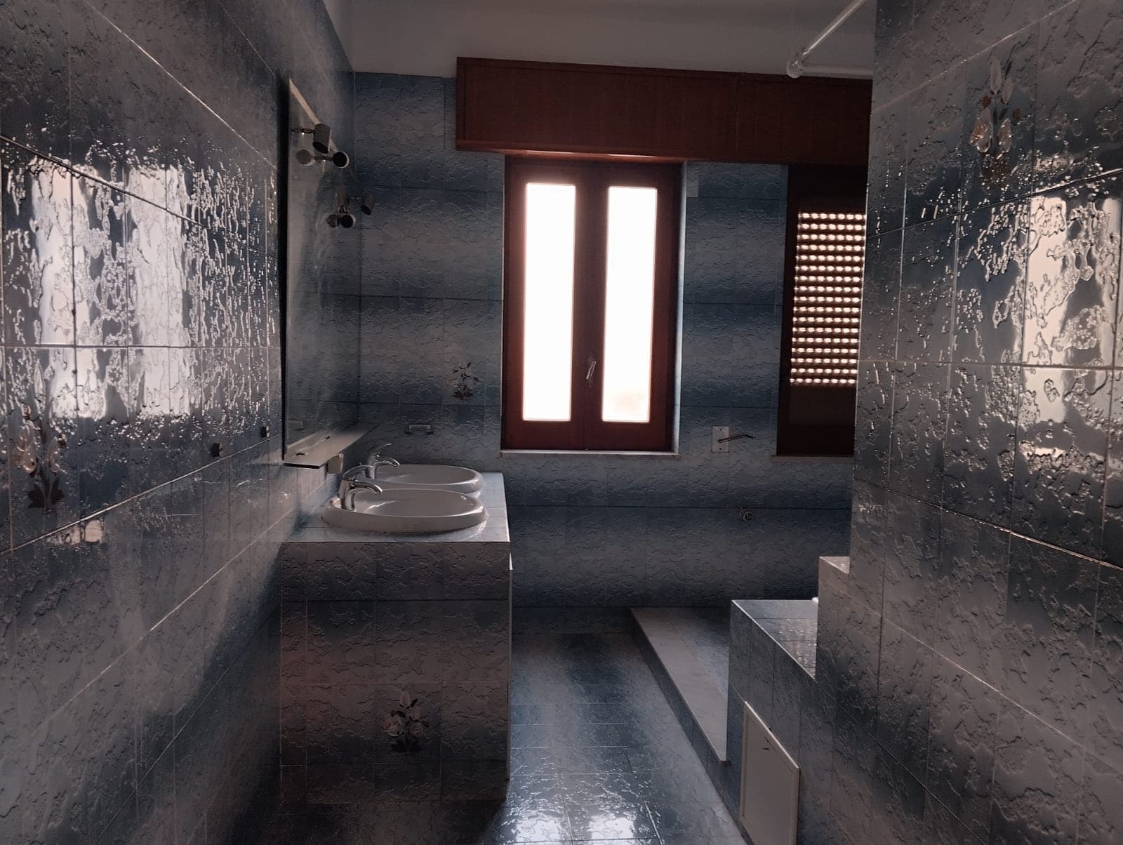 Lavabo doppio in bagno con pareti in piastrelle blu e finestra luminosa, ambiente moderno e funzionale.