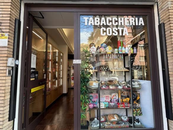 Esterno di una tabaccheria con vetrina che mostra vari prodotti, insegna "Tabaccheria Costa" sopra l'ingresso, ambiente accogliente e ben illuminato, ideale per acquisti di sigarette, gratta e vinci e.
