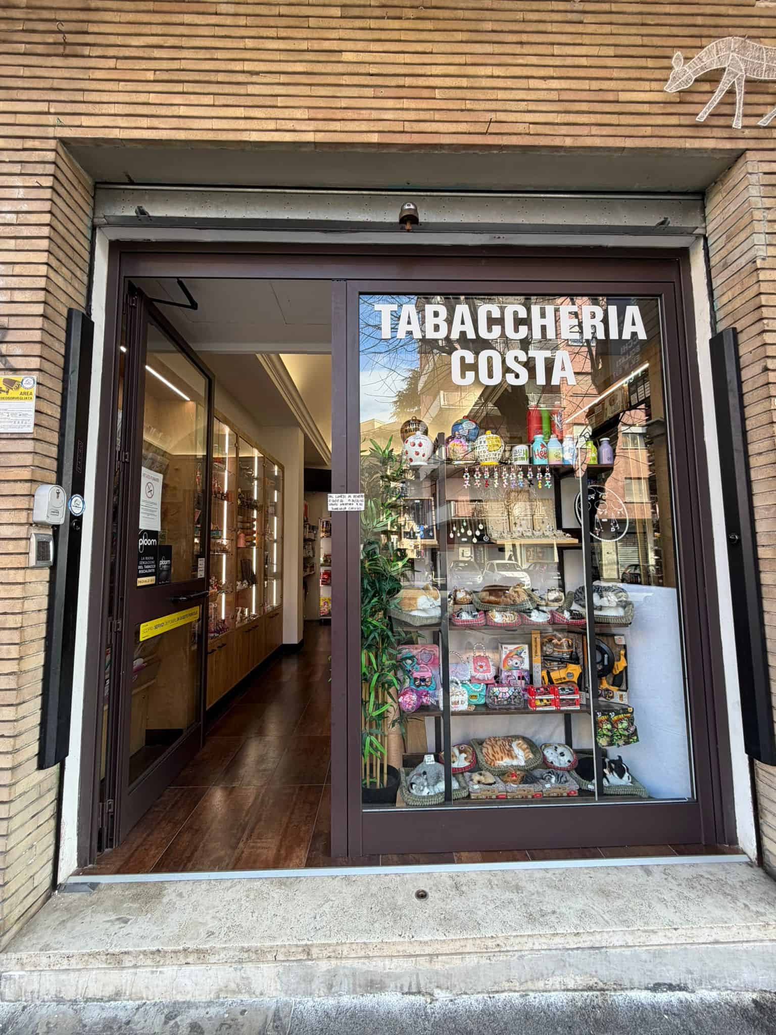 Esterno di una tabaccheria con vetrina che mostra vari prodotti, insegna "Tabaccheria Costa" sopra l'ingresso, ambiente accogliente e ben illuminato, ideale per acquisti di sigarette, gratta e vinci e.