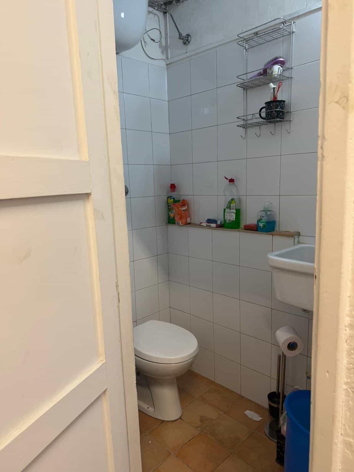 Lavabo e servizi igienici in un bagno compatto con pareti rivestite di piastrelle bianche, scaffali metallici e accessori essenziali, ideale per spazi ridotti.