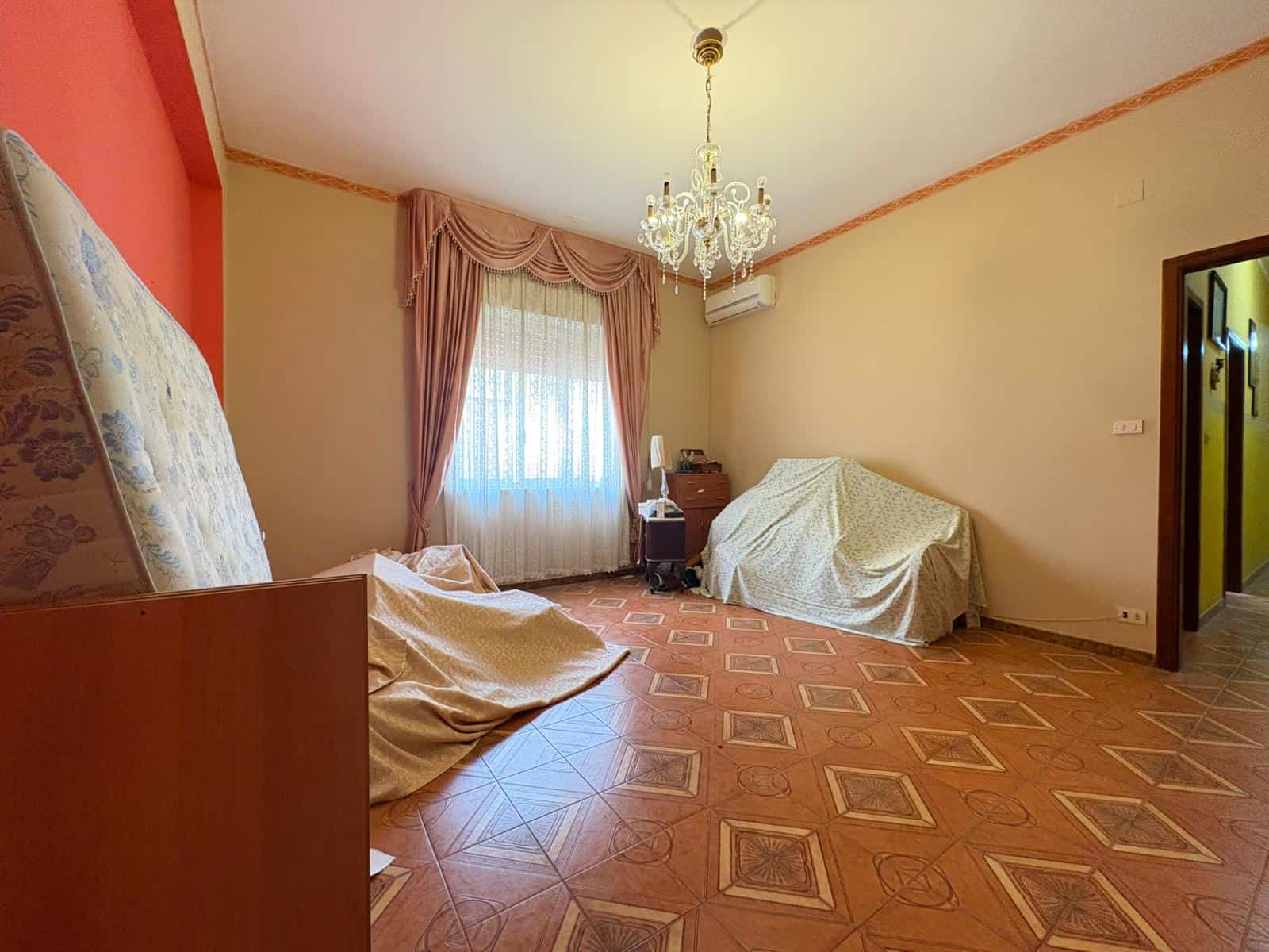Interno di una stanza spaziosa con pavimento in parquet, tende rosa e mobili coperti, illuminata da un lampadario elegante.