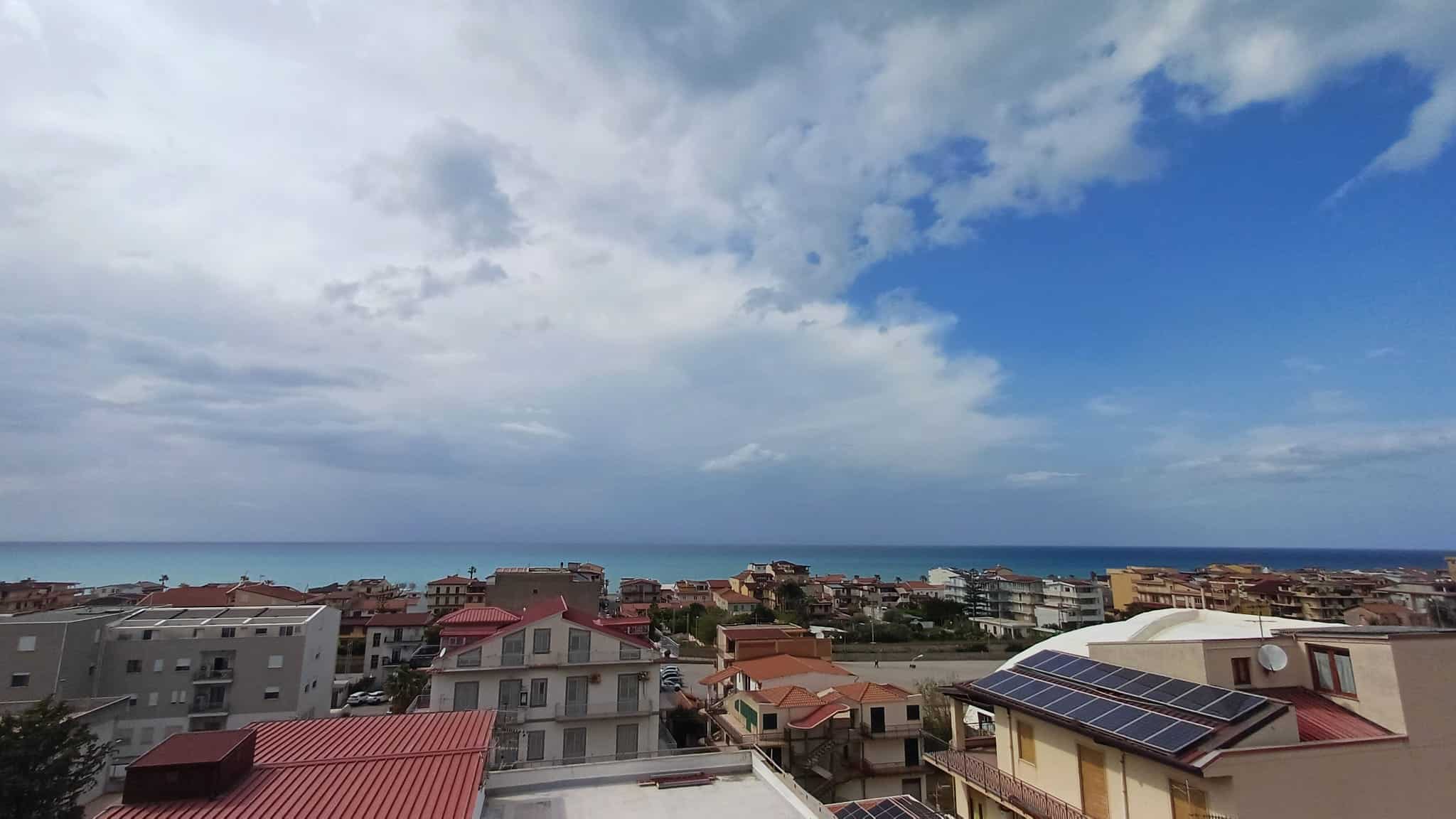 Vista panoramica di case residenziali con tetti solari, sullo sfondo il mare e un cielo nuvoloso, rappresentando un quartiere moderno e sostenibile vicino alla costa.