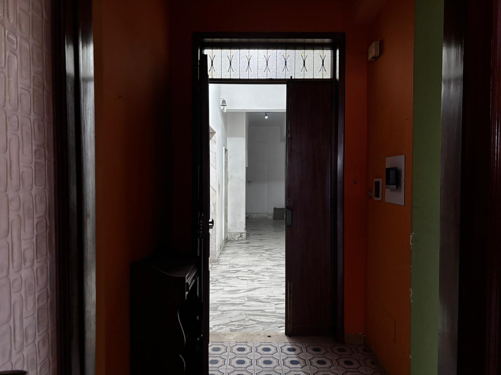 Porta d'ingresso aperta che conduce a un cortile esterno con pavimentazione in pietra, all'interno di un'abitazione con pareti colorate e interruttori moderni, ambiente accogliente e luminoso.