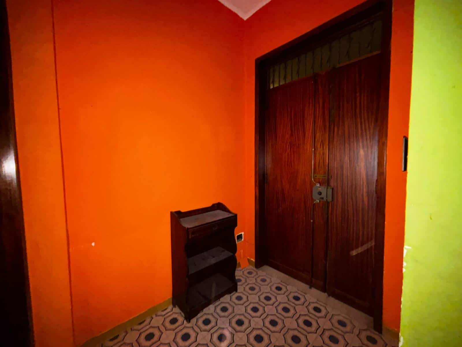 Porte d'ingresso in legno massiccio con serratura di sicurezza, situata in un ingresso colorato con pareti arancioni e verdi, pavimento con piastrelle geometriche.
