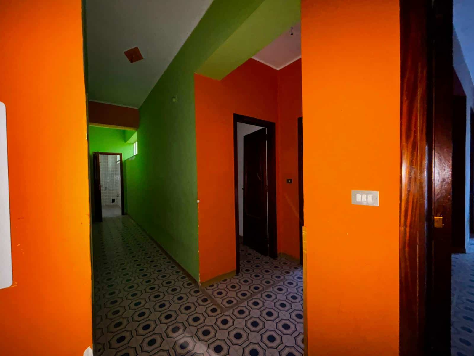 Ampia vista di un corridoio colorato con pareti verdi, arancioni e blu, pavimento con piastrelle geometriche, illuminazione soffusa, ingresso a diverse stanze in un appartamento moderno.