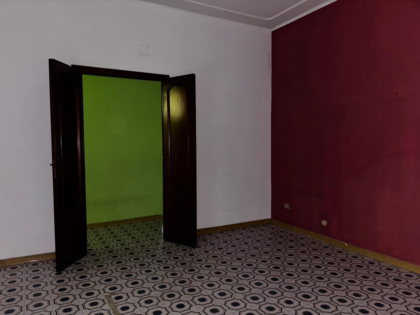Stanza vuota con pareti bianche e una parete color vinaccia, pavimento in piastrelle geometriche, porta aperta verso un'altra stanza verde.
