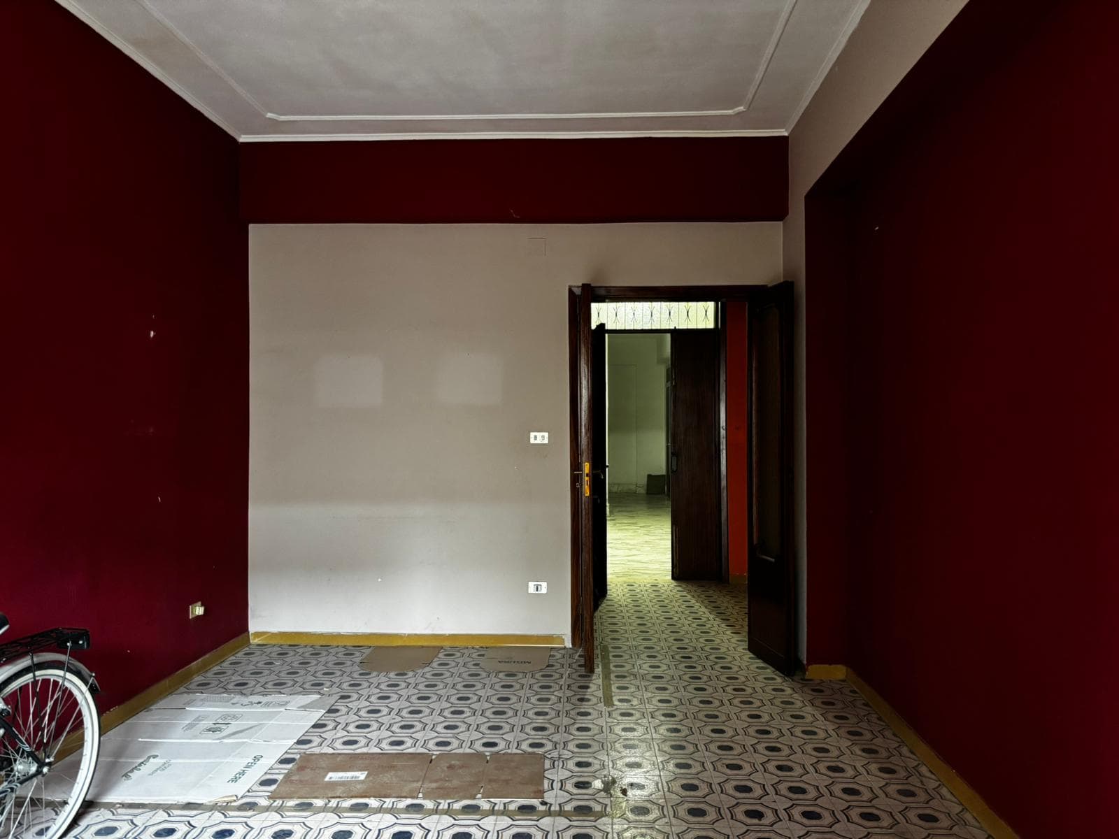 Porte interne in legno in una casa con pareti bordeaux e pavimento in piastrelle decorative, ingresso luminoso con porta aperta che conduce a un'altra stanza.