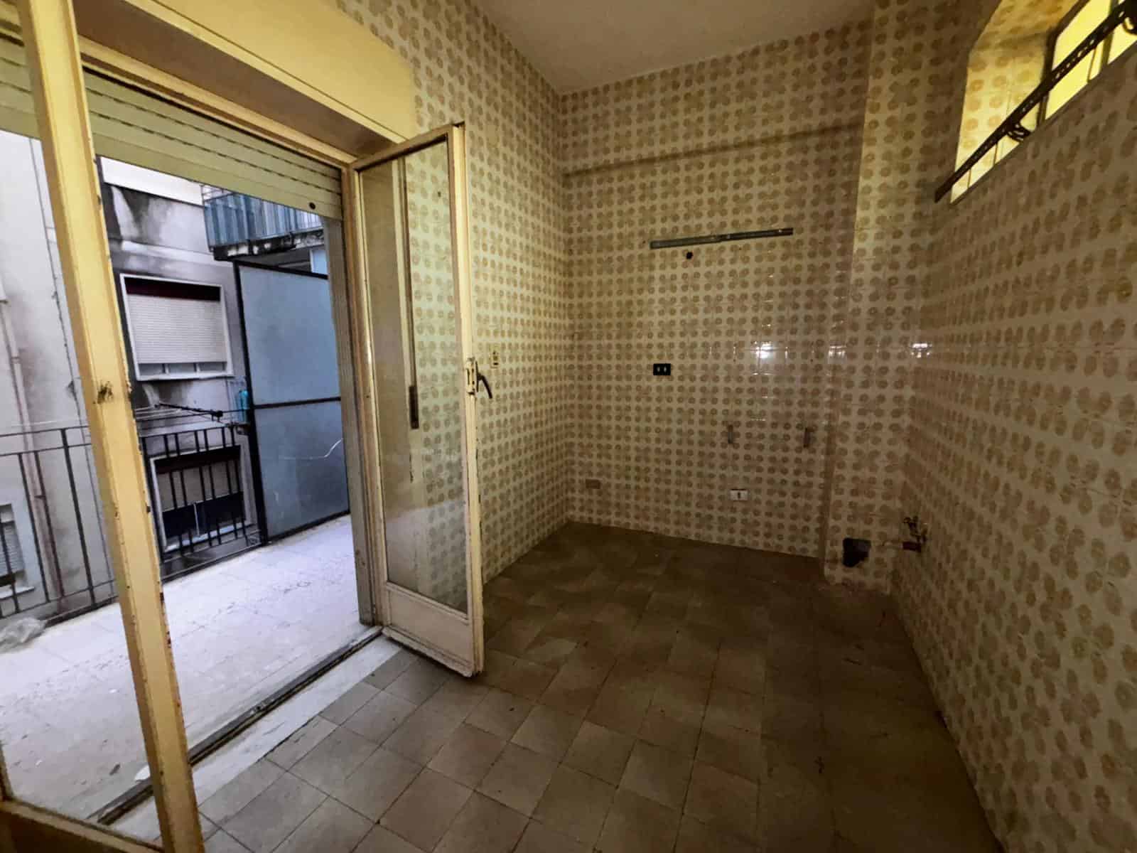 Ampio spazio vuoto in cucina con pareti rivestite di piastrelle beige e accesso a un balcone esterno, ideale per ristrutturazione e personalizzazione.