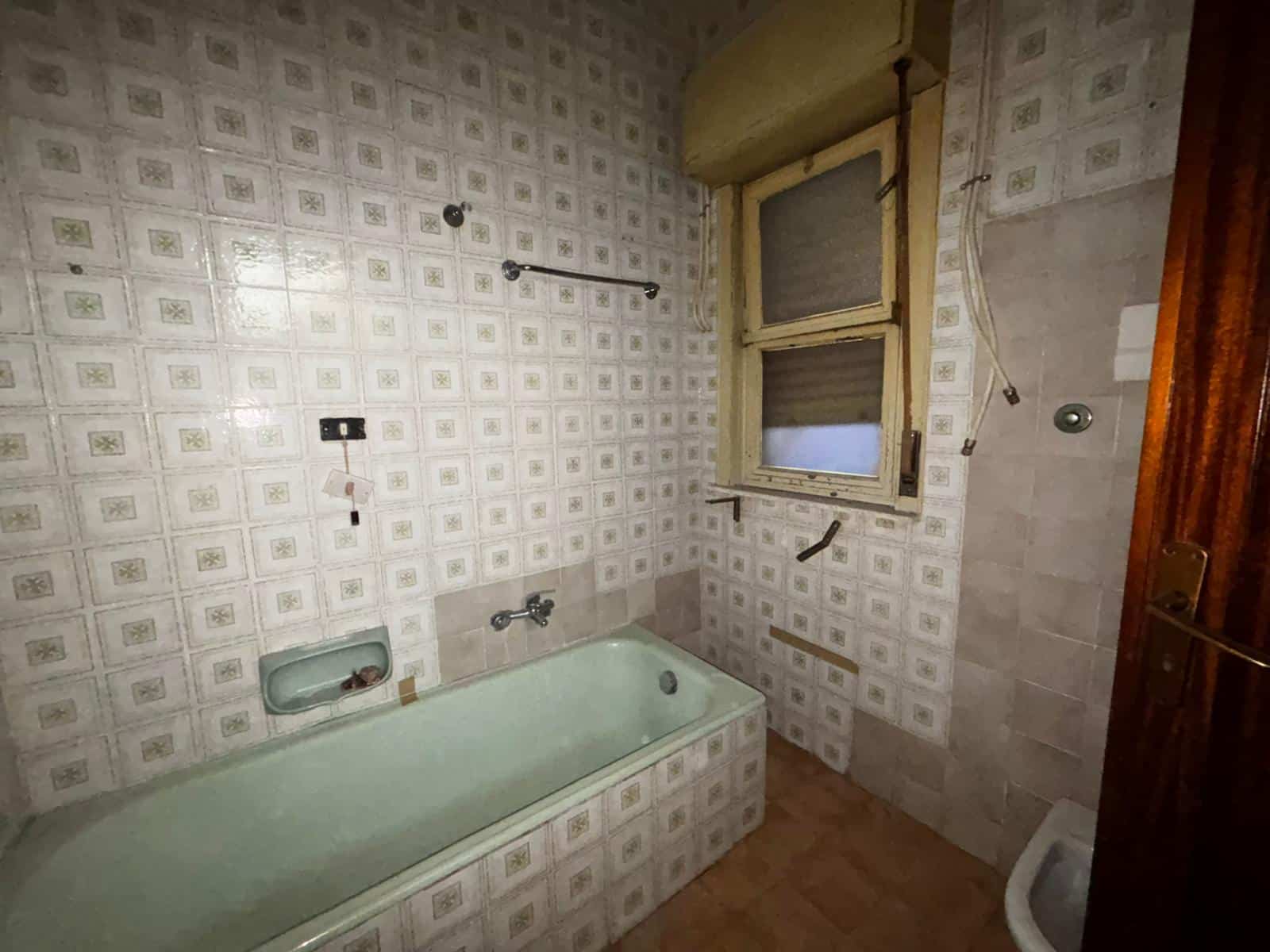 Bagno in fase di ristrutturazione con vasca da bagno, pareti rivestite in piastrelle vintage, finestra con tapparella, elementi di plumbing visibili, ambiente luminoso e spazioso.