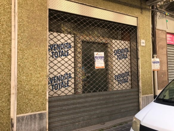 Vendita locale commerciale chiuso con grate metalliche e cartelli "VENDITA" affissi, situato in zona urbana con veicolo bianco parcheggiato di fronte.