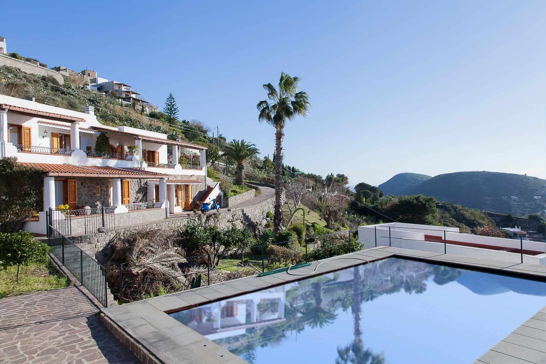 Piscina all'aperto con vista panoramica sulle colline e palme, case in stile mediterraneo, ambiente tranquillo e soleggiato, ideale per relax e vacanze in Toscana.