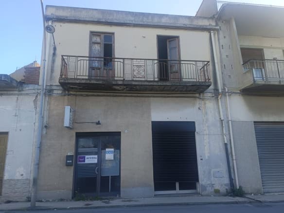 Balcone in ferro battuto su edificio storico in centro città, con finestre e porta d'ingresso al piano terra, in un contesto urbano con edifici residenziali e commerciali.
