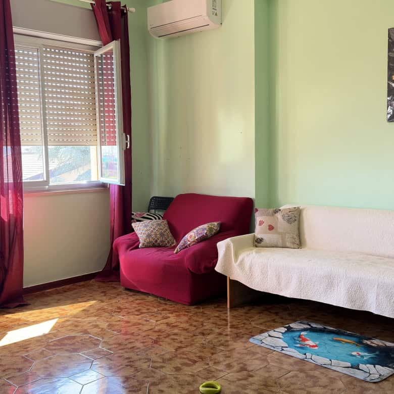 Ampia zona living con divano e poltrona, luminosa grazie alle grandi finestre con tende rosse, pareti verde pastello e decorazioni moderne, ideale per ambienti accoglienti e di design.