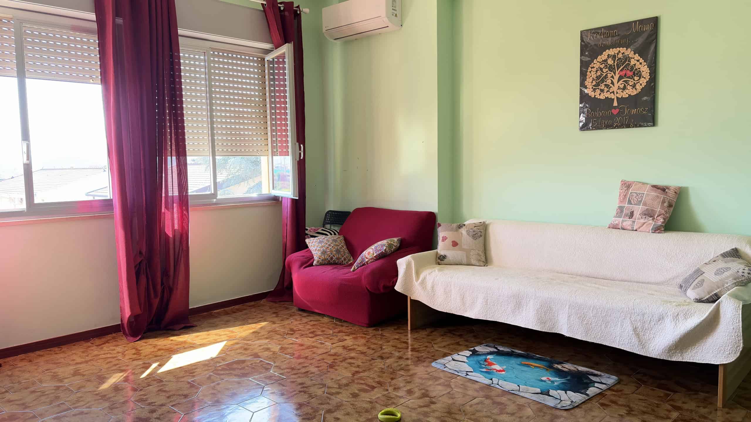 Ampia zona living con divano e poltrona, luminosa grazie alle grandi finestre con tende rosse, pareti verde pastello e decorazioni moderne, ideale per ambienti accoglienti e di design.