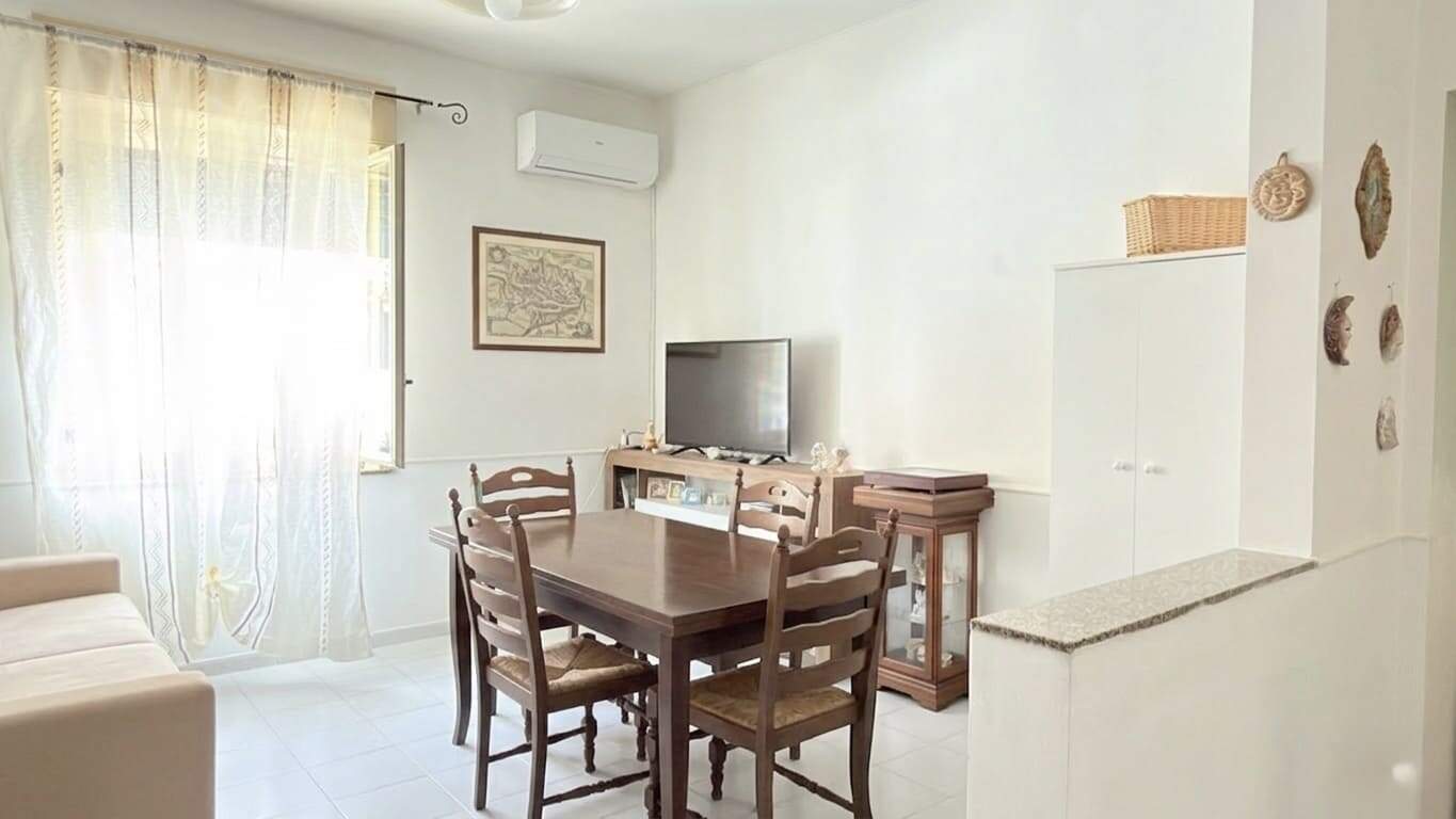 Appartamento luminoso con soggiorno e zona pranzo, arredato in stile semplice e funzionale, con accesso a una finestra con tende leggere, ideale per affitti turistici o residenziali.