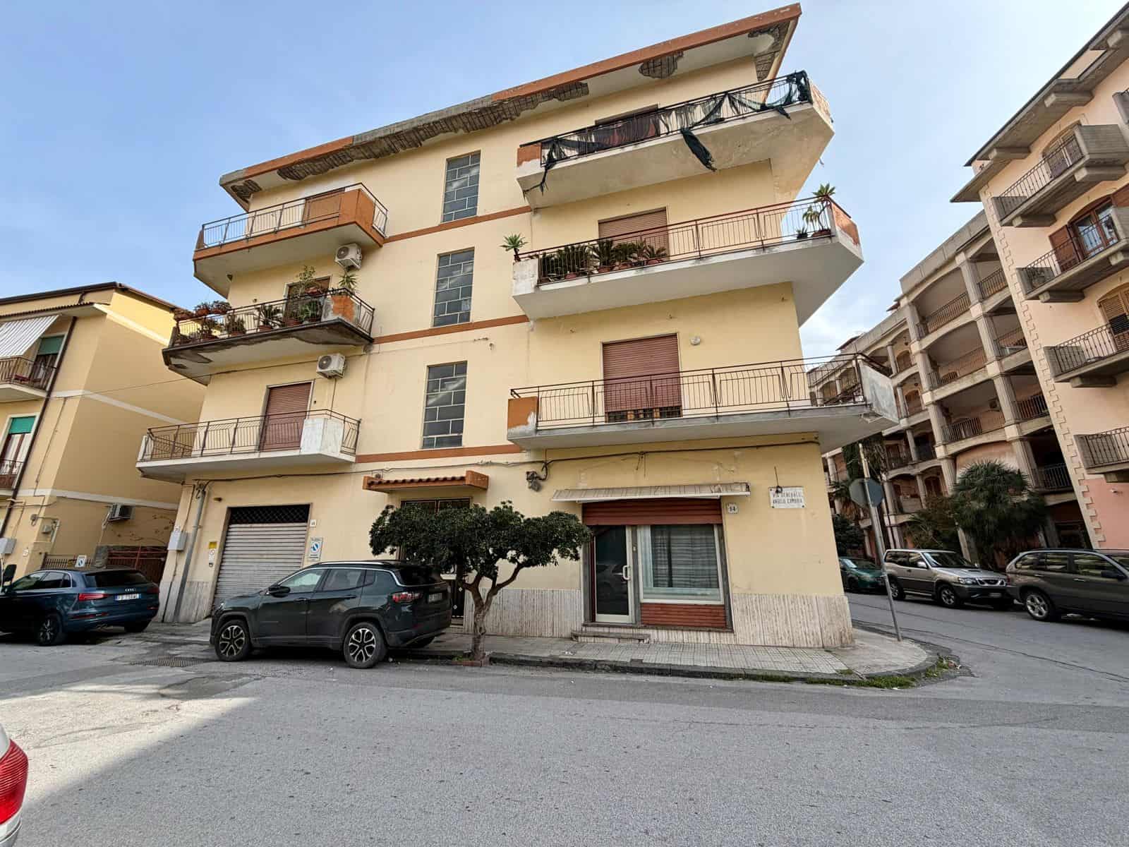 Balcone e facciata di un edificio residenziale in zona urbana, con auto parcheggiate davanti, piante e balconi fioriti, in stile mediterraneo, in una giornata soleggiata.
