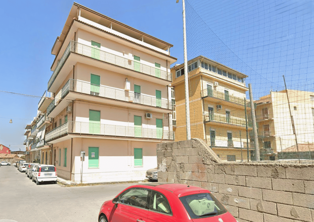 Appartamenti residenziali in edificio multifamiliare con balconi e finestre verdi, situato in zona urbana soleggiata e tranquilla, con auto parcheggiate lungo la strada e rete di recinzione intorno al.