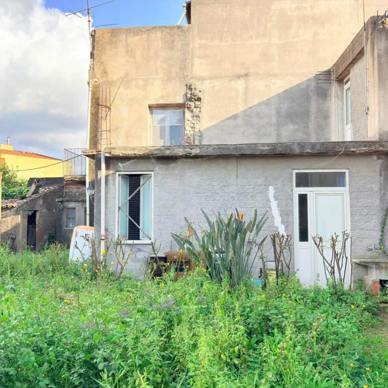 Veranda di una casa in stile rustico con giardino verde e piante, vista esterna di un'abitazione in zona urbana, con pareti in pietra e finestre moderne, ambiente tranquillo e naturale.