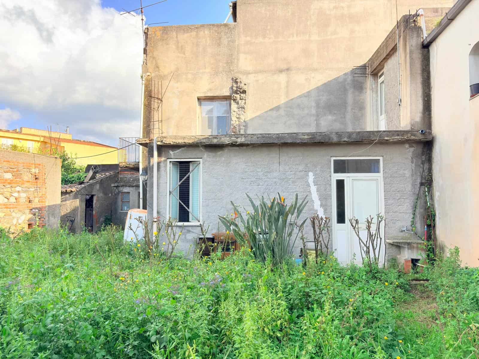 Veranda di una casa in stile rustico con giardino verde e piante, vista esterna di un'abitazione in zona urbana, con pareti in pietra e finestre moderne, ambiente tranquillo e naturale.