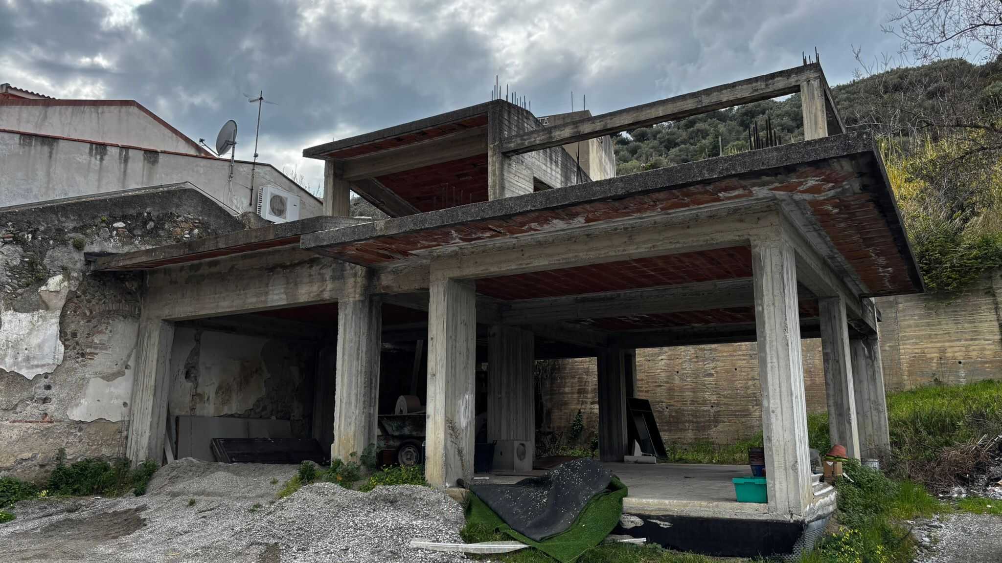 Vicolo di un edificio in costruzione con struttura in cemento armato, ambiente esterno con terreno non rifinito e cielo nuvoloso, in un'area in fase di sviluppo edilizio.